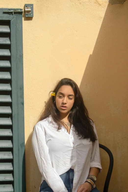 a woman sitting on a chair in front of a building, an album cover, by Alexis Grimou, pexels contest winner, hyperrealism, wearing a white button up shirt, with yellow flowers around it, headshot profile picture, pale-skinned persian girl