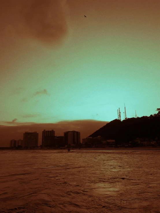 a large body of water with buildings in the background, a matte painting, inspired by Elsa Bleda, unsplash, brutalism, on the beach during sunset, brown and cyan color scheme, grainy photograph, rio de janeiro