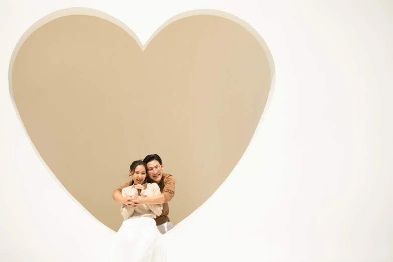 a man and a woman hugging in front of a heart, a picture, by Tan Ting-pho, 8k studio photography, plain walls |light hearted, beige, white hue