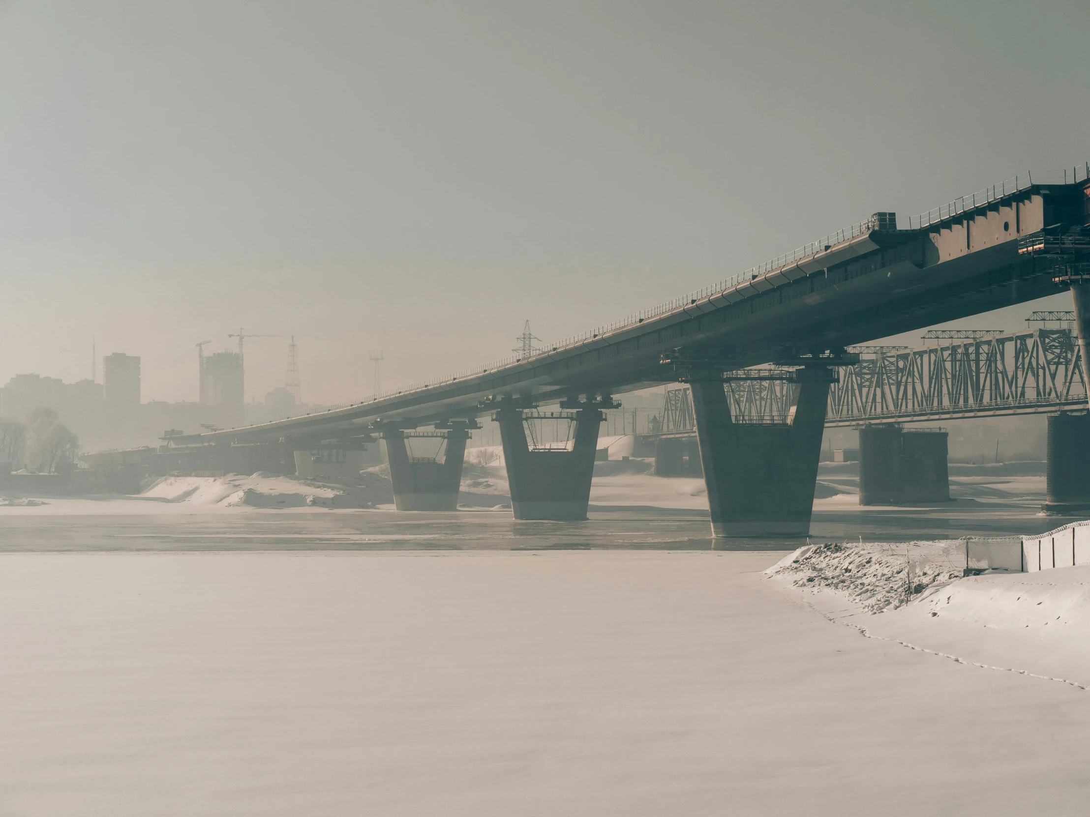 a train traveling over a bridge over a river, inspired by Elsa Bleda, pexels contest winner, romanticism, norilsk, gray fog, ground covered with snow, tall bridge with city on top