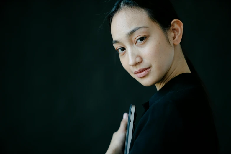 a close up of a woman holding a cell phone, a character portrait, inspired by Qian Xuan, pexels contest winner, standing with a black background, kiko mizuhara, ethnicity : japanese, wearing a black shirt