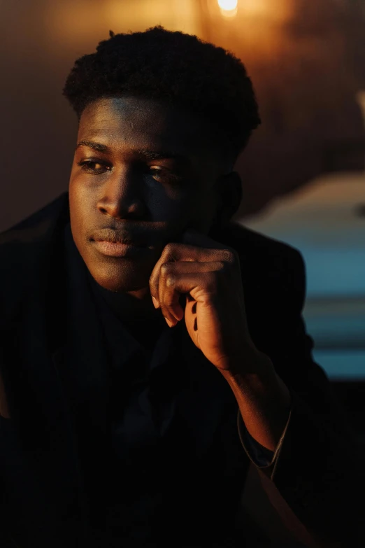 a man sitting in front of a car talking on a cell phone, an album cover, by Maxwell Bates, trending on pexels, renaissance, young man with beautiful face, ( ( dark skin ) ), portrait soft light, androgynous person