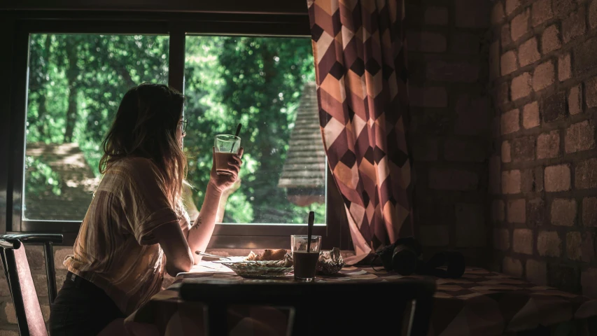 a woman sitting at a table with a glass of wine, a picture, inspired by Elsa Bleda, pexels contest winner, looking out open window, in a tree house, sitting on a mocha-colored table, green light coming from window