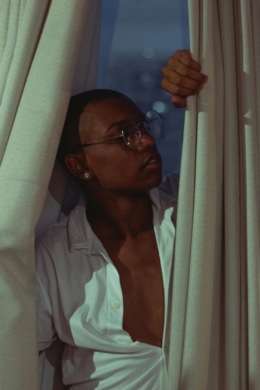 a man in a white shirt looking out of a window, an album cover, inspired by Terrell James, trending on pexels, with square glasses, androgynous person, fully covered in drapes, summer night