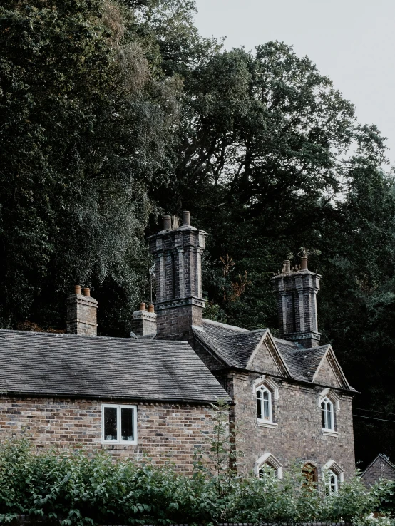 a couple of people standing in front of a building, by IAN SPRIGGS, pexels contest winner, several cottages, chimney, with dark trees in foreground, staggered terraces