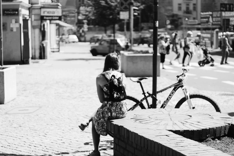 a woman sitting on a bench next to a bike, a black and white photo, summer street, with a backpack, wearing in a summer dress, looking from behind