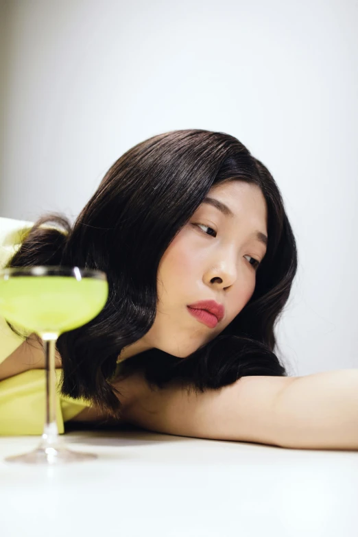 a woman sitting at a table with a glass of wine, an album cover, inspired by Fei Danxu, holding a martini, jaeyeon nam, looking away from camera, full of greenish liquid