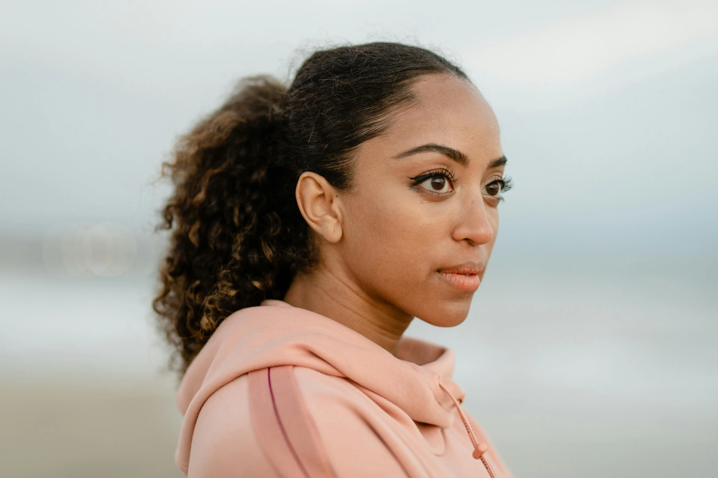 a woman standing on top of a beach next to the ocean, a character portrait, by Lily Delissa Joseph, pexels contest winner, wearing a pink hoodie, portrait of vanessa morgan, close up half body shot, with a ponytail