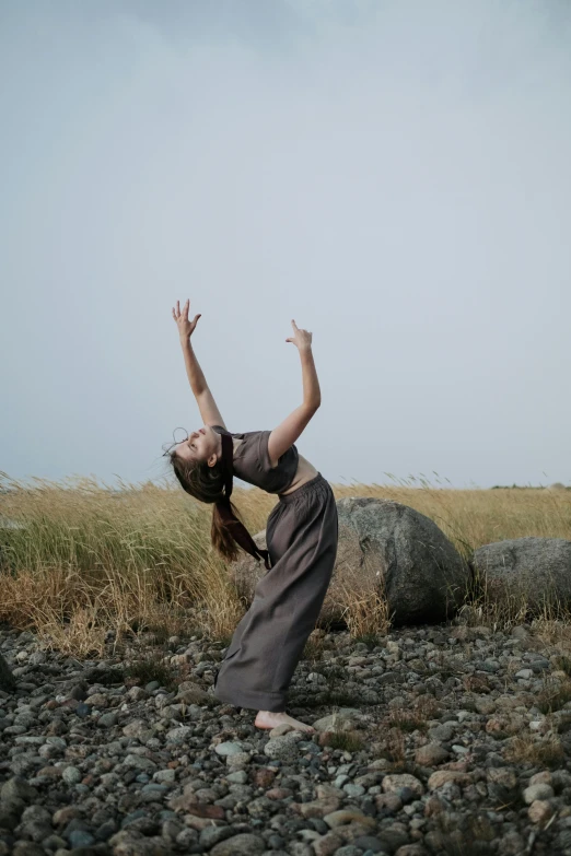 a woman standing on top of a rocky beach, an album cover, by Jessie Algie, land art, contemporary dance poses, in a grassy field, crying and reaching with her arm, late summer evening