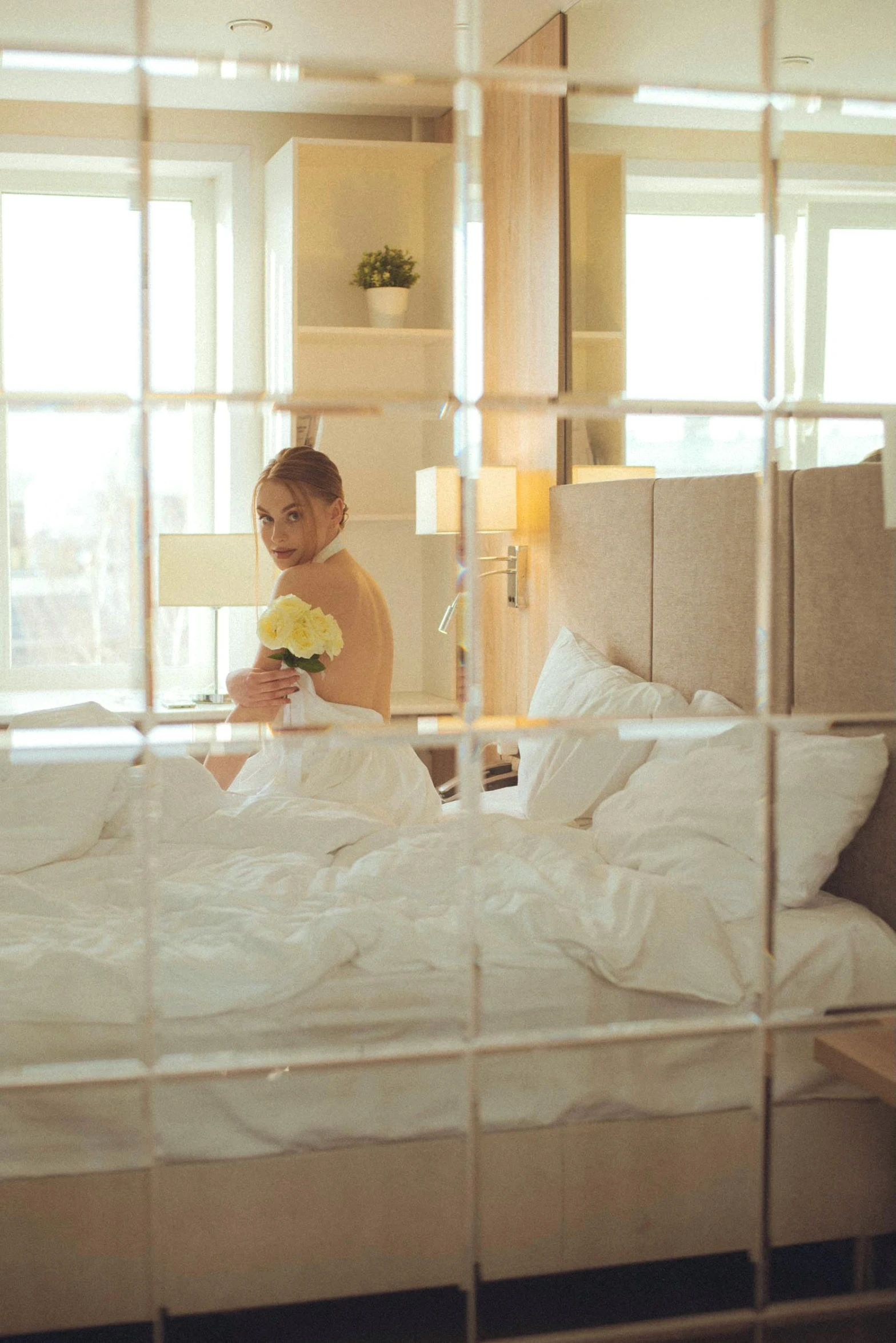 a woman sitting on top of a bed next to a window, holding flowers, hotel room, wearing translucent sheet, mirrored