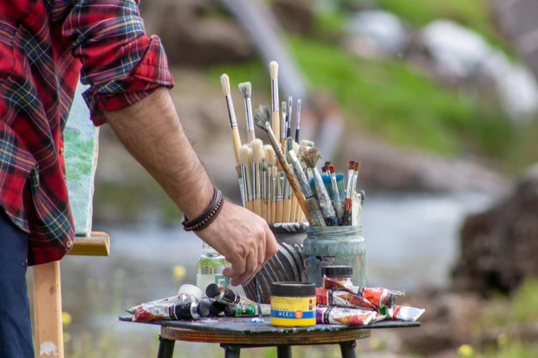 a man standing next to a table filled with art supplies, inspired by Stanhope Forbes, pexels contest winner, outdoor scene, holding paintbrushes, al fresco, environmental shot