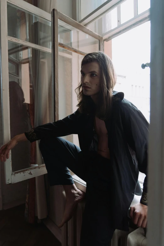 a man with long hair sitting on a window sill, an album cover, unsplash, androgynous male, at a fashion shoot, in doors, steven klein