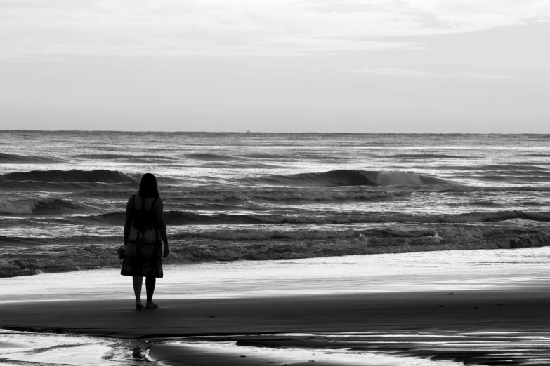 a woman standing on top of a beach next to the ocean, a black and white photo, by Caro Niederer, unsplash, the sea of sadness, end of the day, busy but lonely, woman in black robes