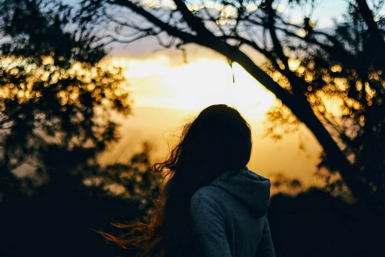 a woman standing in front of a tree at sunset, pexels contest winner, girl wearing hoodie, profile image, back - shot, looking onto the horizon