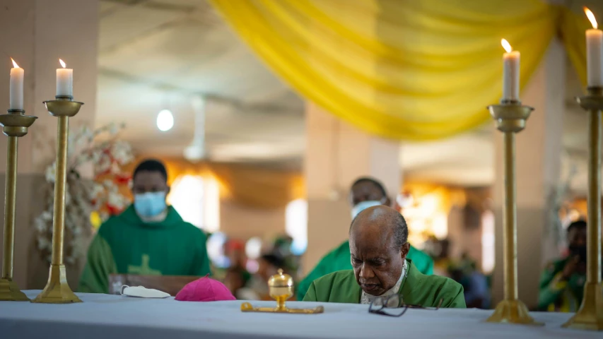 a man sitting at a table with candles in front of him, draped in pink and green robes, emmanuel shiru, inauguration, thumbnail