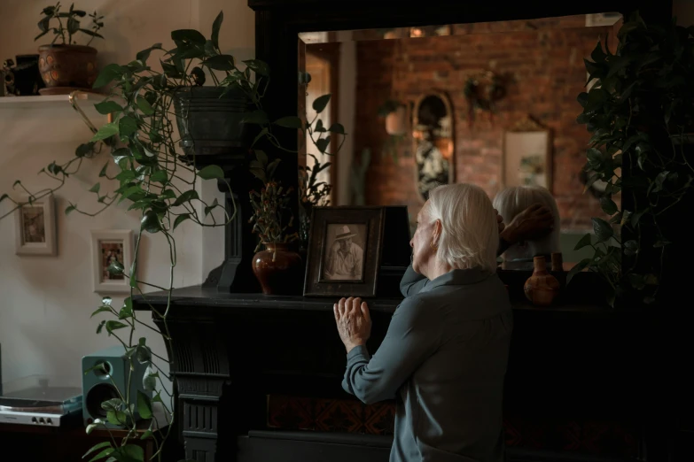 a woman standing in front of a mirror in a living room, a picture, by Everett Warner, pexels contest winner, mourning family, white haired lady, old home decor, in front of a fireplace