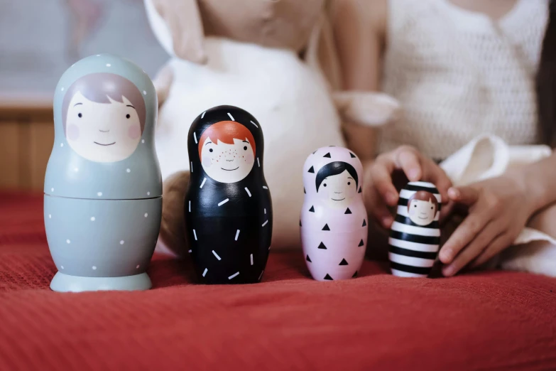 a woman sitting on a bed next to three nesting dolls, unsplash, sarah andersen, tiny stars, on wooden table, siblings