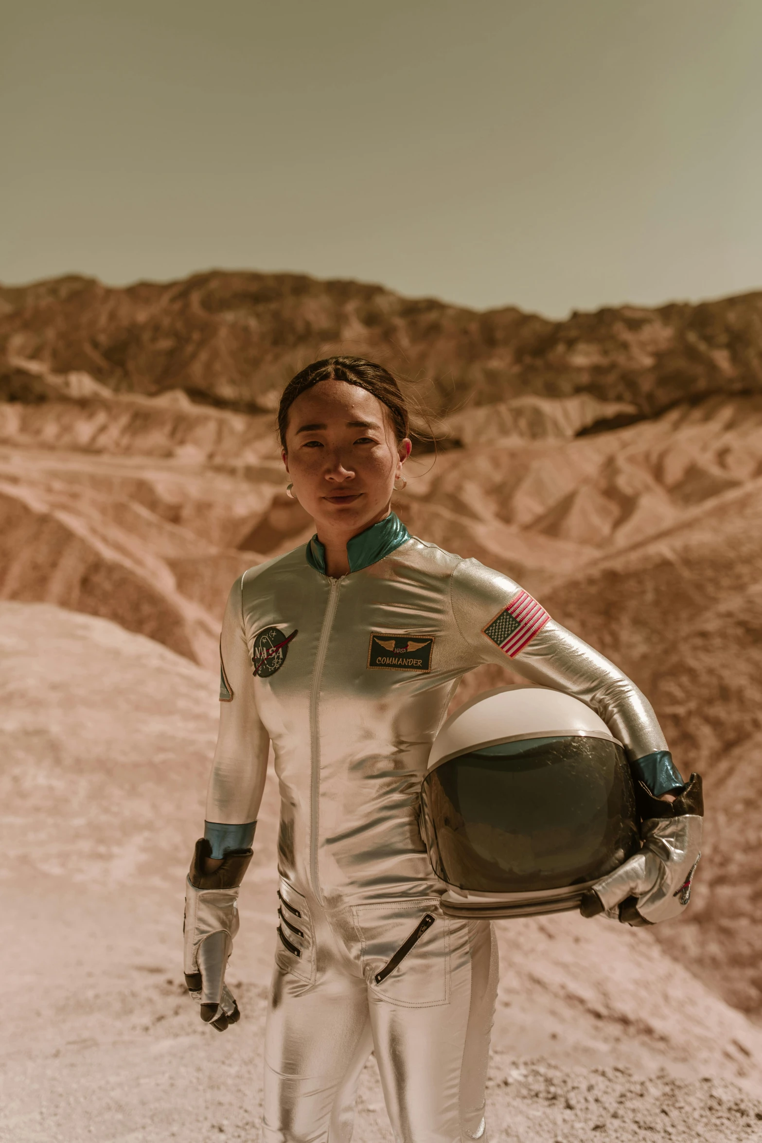 a man in a space suit holding a helmet, jamie chung, standing in a desert, louise zhang, flight suit and gloves