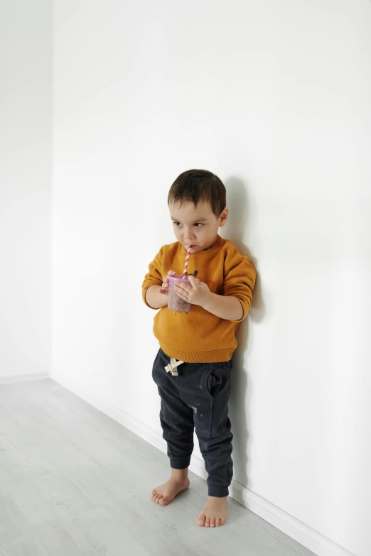 a young boy leaning against a wall using a cell phone, a picture, inspired by Sarah Lucas, pexels, in front of white back drop, drinking cough syrup, brown pants, toddler