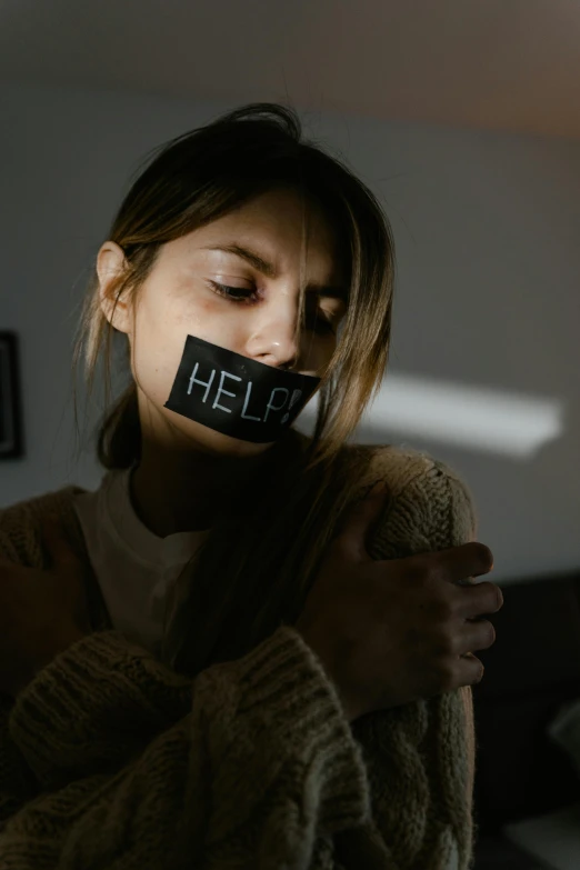 a woman holding a piece of tape over her mouth, a picture, inspired by Elsa Bleda, trending on pexels, antipodeans, woman holding sign, help, low key light, a labeled
