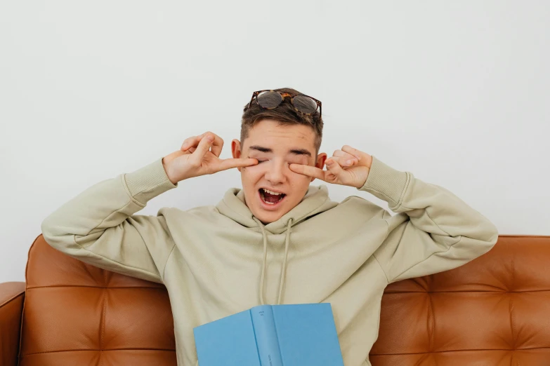 a young man sitting on top of a couch holding a book, trending on pexels, holding his hands up to his face, tommy 1 6 years old, goggles on forehead, wearing a tracksuit