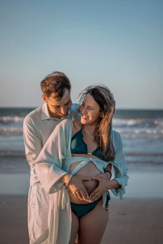 a man standing next to a woman on a beach, a colorized photo, by Gwen Barnard, unsplash, maternity feeling, wearing simple robes, beautifully soft lit, birth