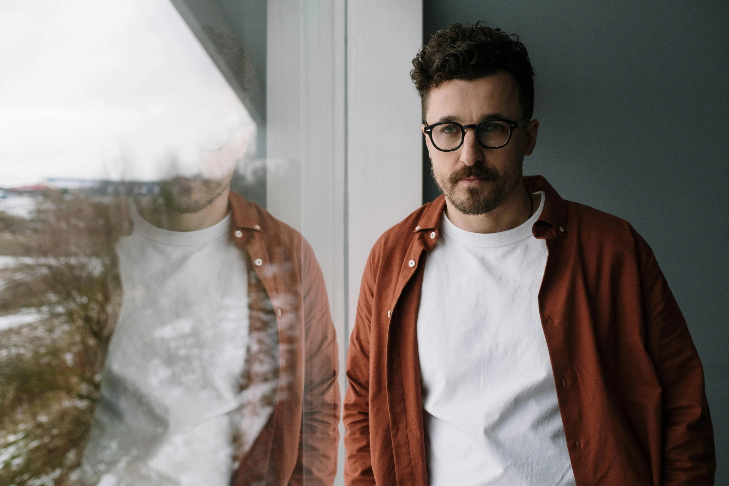 a man standing in front of a window, an album cover, inspired by Dan Smith, pexels contest winner, wearing a white button up shirt, man with glasses, asher duran, grainy photo