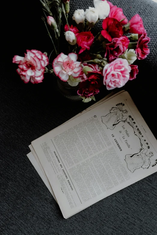 a cup of coffee sitting on top of a table next to a book, holding flowers, early 1900s newspaper, on black paper, pink flowers