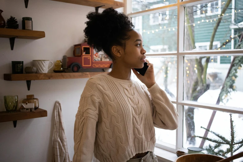 a woman standing in front of a window talking on a cell phone, by Lily Delissa Joseph, trending on pexels, happening, natural hair, wearing a white sweater, calmly conversing 8k, cute girl