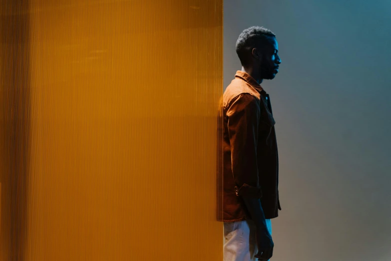 a man standing in front of a yellow wall, by Clifford Ross, pexels contest winner, dark skinned, light lighting side view, orange and blue tones, relaxed. gold background