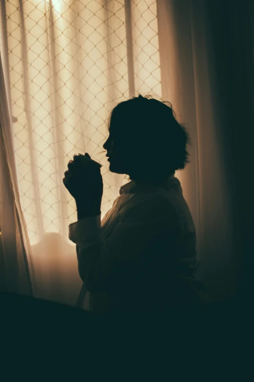 a silhouette of a woman sitting in front of a window, inspired by Elsa Bleda, pexels, praying, hand on her chin, morning coffee, dark hazy room