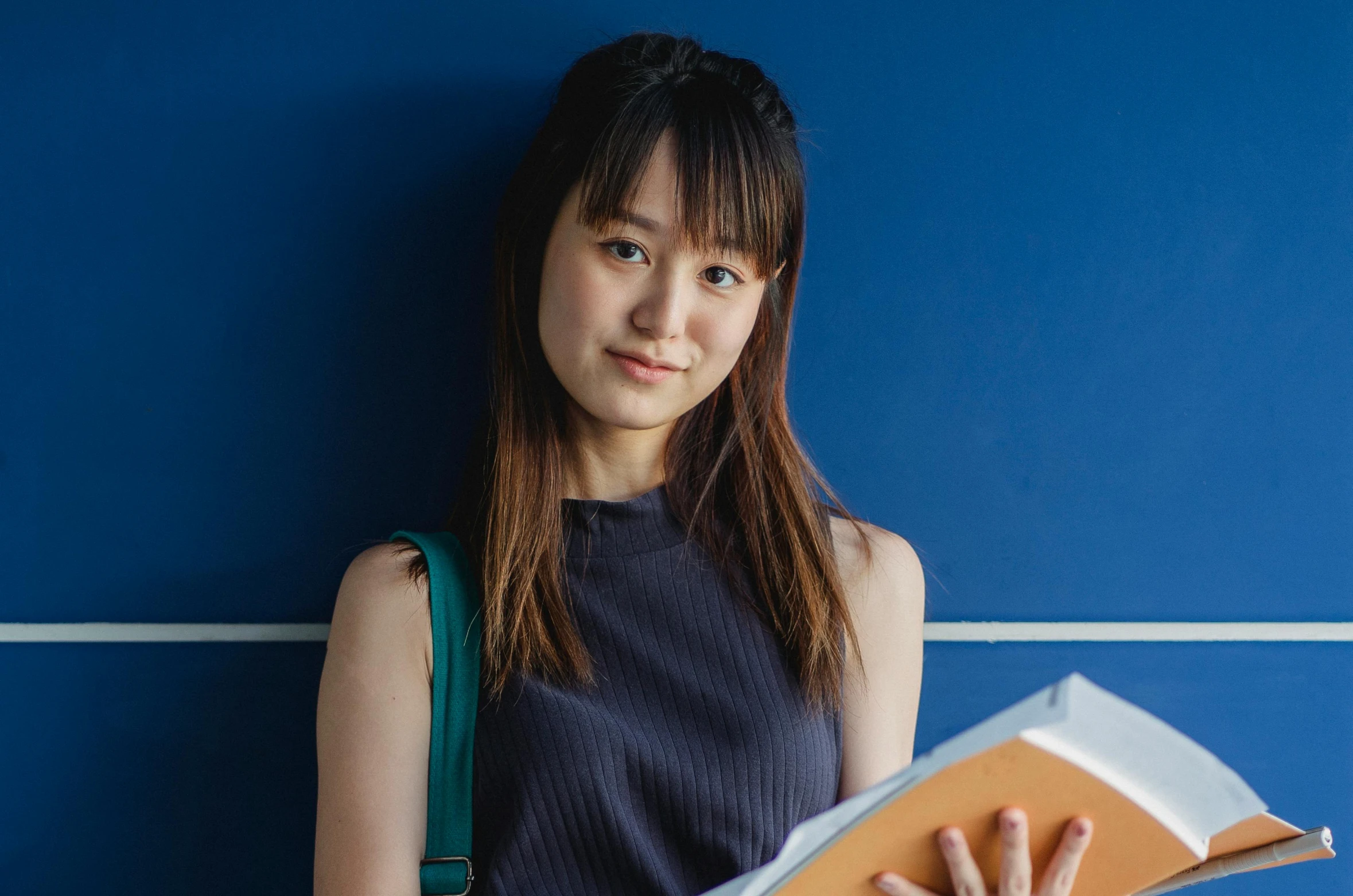 a woman standing in front of a blue wall holding a book, inspired by Ma Yuanyu, pexels contest winner, shin hanga, she is wearing a black tank top, avatar image, studious, ethnicity : japanese