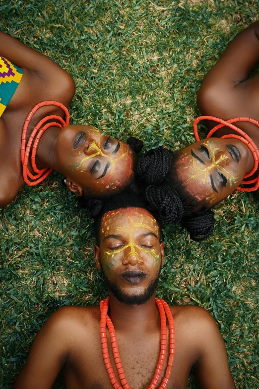 a group of people laying on top of a lush green field, an album cover, inspired by Ras Akyem, pexels contest winner, afrofuturism, tribal face paintings, three heads, view from above, elaborate patterned makeup