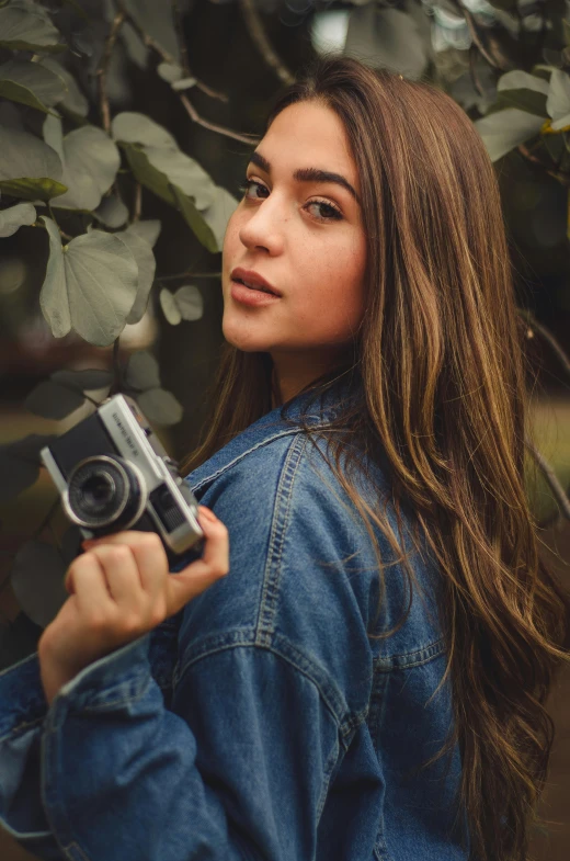 a woman holding a camera in front of a tree, by Robbie Trevino, ana de armas portrait, teen girl, high quality image, icon