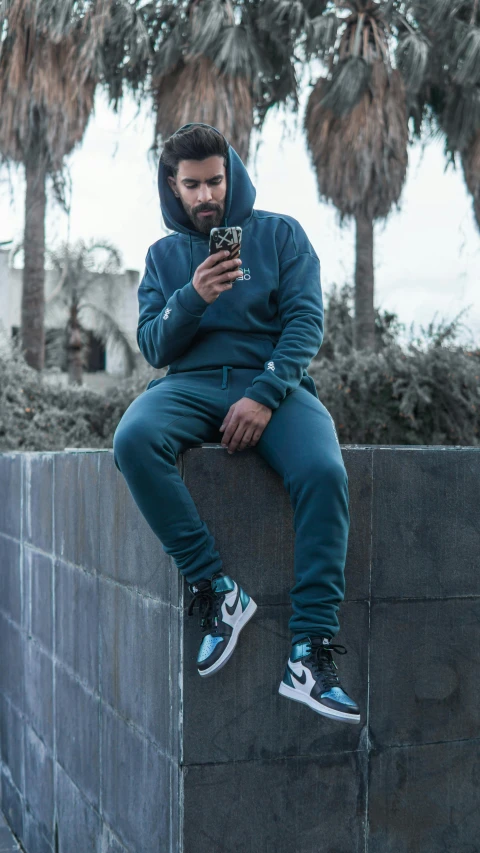 a man sitting on a wall looking at his cell phone, an album cover, by Robbie Trevino, trending on pexels, renaissance, teal uniform, wearing a track suit, full body photo, navy blue