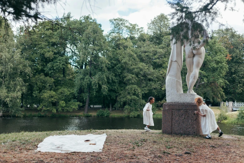 a couple of people standing next to a statue, a statue, by Emma Andijewska, temporary art, in a park and next to a lake, wearing torn white cape, standing over a tomb stone, ignant