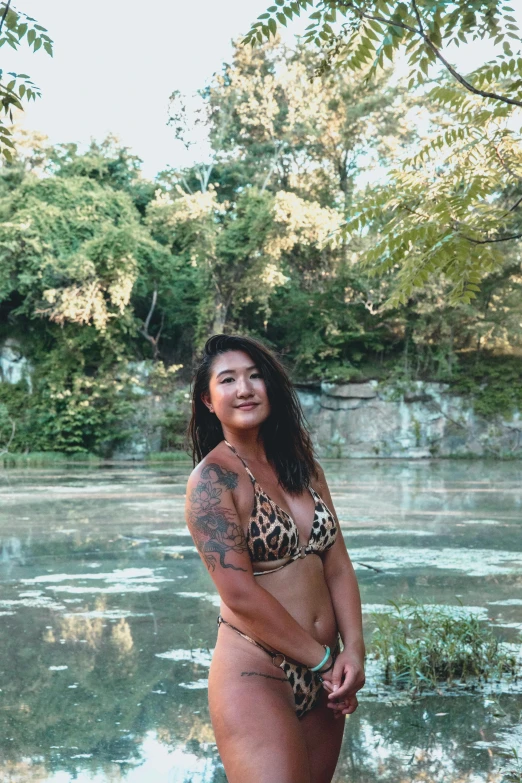 a woman in a bikini standing next to a body of water, of taiwanese girl with tattoos, lush surroundings, & a river, profile image