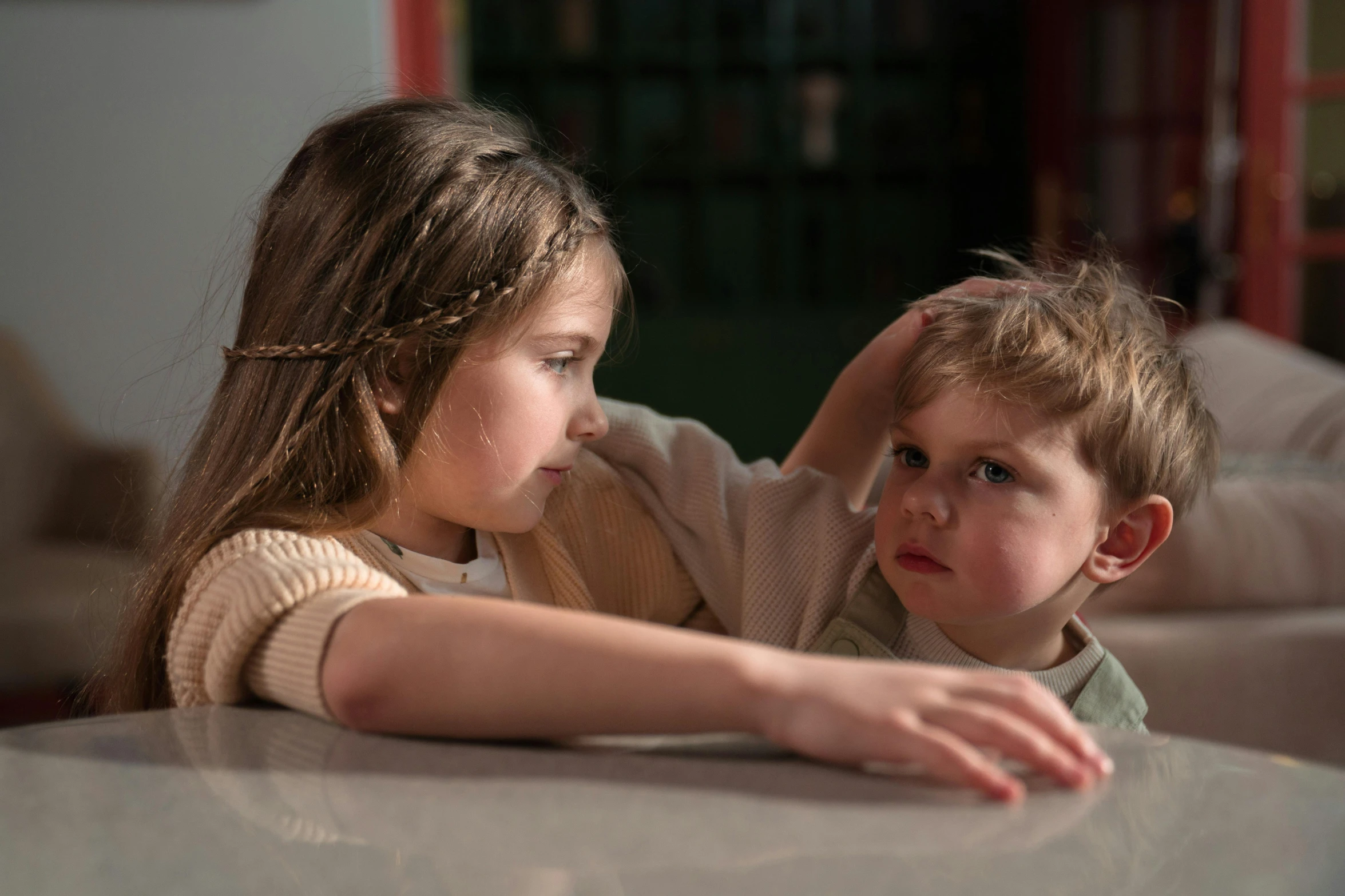 a boy and a girl sitting at a table, by Sylvia Wishart, pexels, realism, scratching head, little brother, soft light.4k, looking away