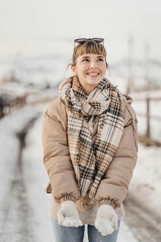 a woman standing on the side of a road in the snow, trending on pexels, beige, with a happy expression, tartan scarf, modest