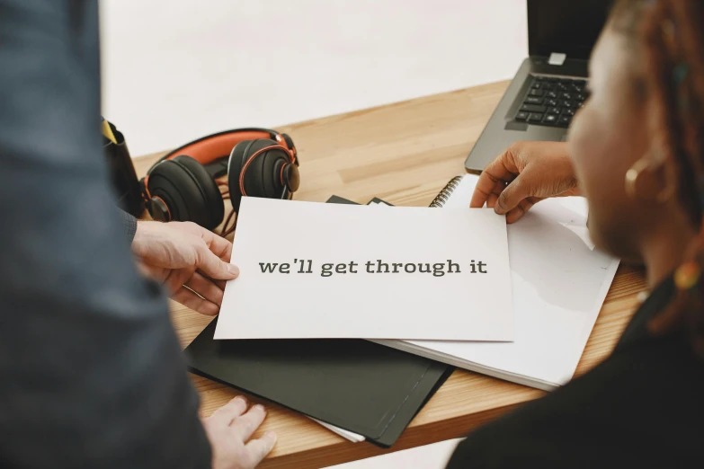 a person holding a piece of paper that says we'll get through it, by Julia Pishtar, trending on unsplash, private press, in an call centre office, on a white table, 9 9 designs, a wooden