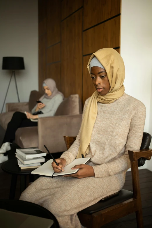 a woman sitting on a chair in a living room, inspired by Maryam Hashemi, trending on pexels, hurufiyya, medium shot of two characters, writing in journal, wearing a head scarf, serious business