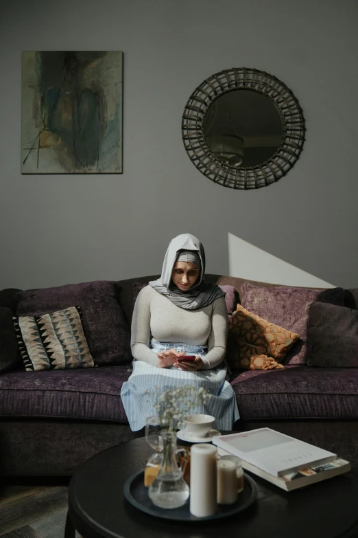 a woman sitting on a couch in a living room, a portrait, inspired by Maryam Hashemi, pexels contest winner, hurufiyya, white hijab, grey, seated at a table, cinematic full body shot