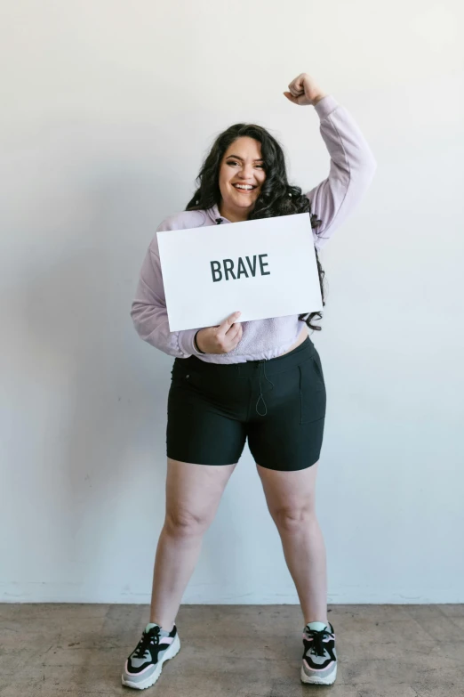 a woman holding a sign that says brave, pexels contest winner, curvy body, wearing shorts, hero pose, kailee mandel