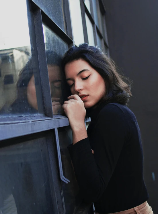 a woman leaning against a window talking on a cell phone, a picture, inspired by Elsa Bleda, trending on unsplash, realism, windblown dark hair, hailee steinfeld, her eyes are closed, wearing a black sweater