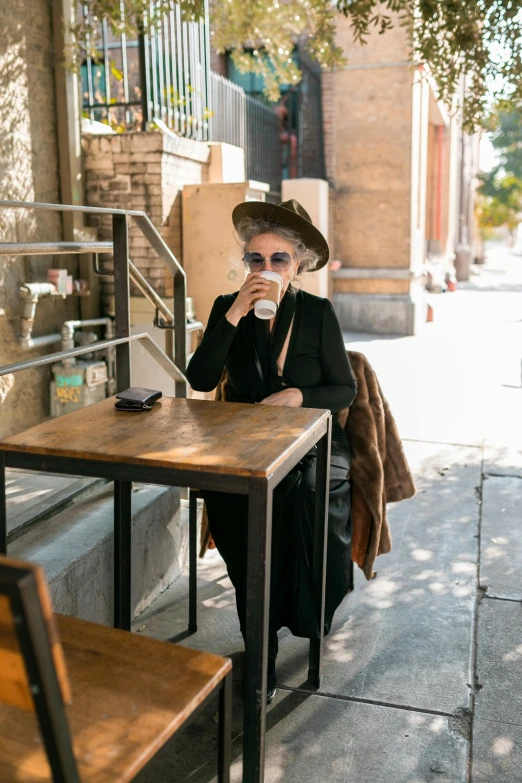 a woman sitting at a table with a cup of coffee, wearing black old dress and hat, tehran, on sidewalk, profile image