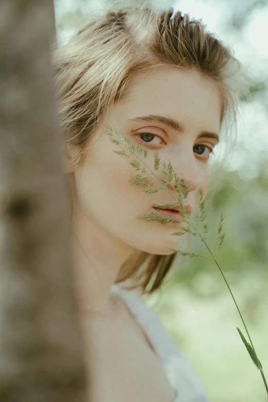 a woman standing next to a tree holding a flower, inspired by Elsa Bleda, aestheticism, prominent cheekbones, hiding in grass, portrait featured on unsplash, portrait image