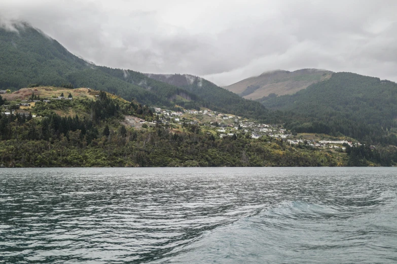 a body of water with mountains in the background, a picture, by Hazel Armour, unsplash, hurufiyya, in the foreground a small town, the photo was taken from a boat, overcast, kahikatea