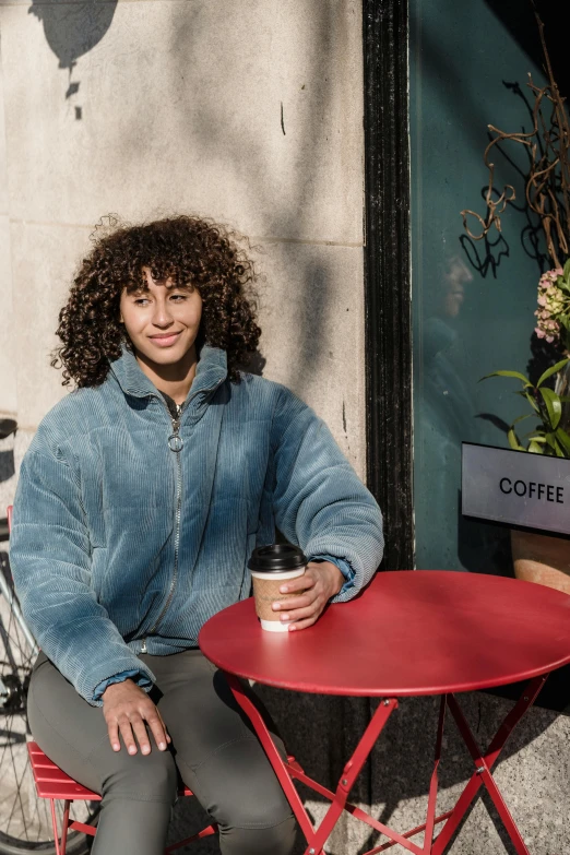 a woman sitting at a table with a cup of coffee, curly haired, wearing a blue hoodie, in london, wearing a jeans jackets