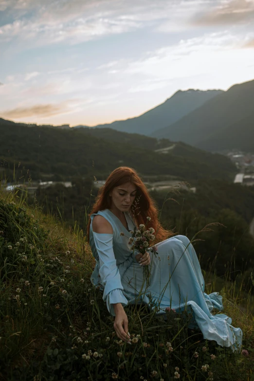 a woman in a blue dress sitting on a hill, inspired by Elsa Bleda, trending on pexels, alaska, maiden with copper hair, early morning, holding flowers