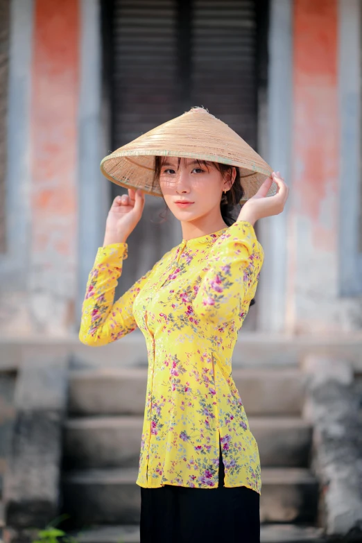 a woman standing in front of a building with a hat on her head, a picture, inspired by Tan Ting-pho, renaissance, wearing a yellow dress, vietnam, upper body image, square